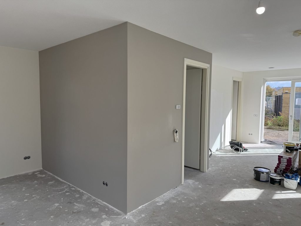 Woonkamer van een net opgeleverde nieuwbouwwoning met een grijs gesausde wand en verfemmers op de betonvloer